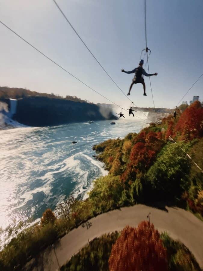 Simulated scene from Niagara Takes Flight showing riders soaring along ziplines above the Niagara River gorge during peak fall foliage, with Horseshoe Falls visible in the background and brilliant red and orange trees lining the curved path below.