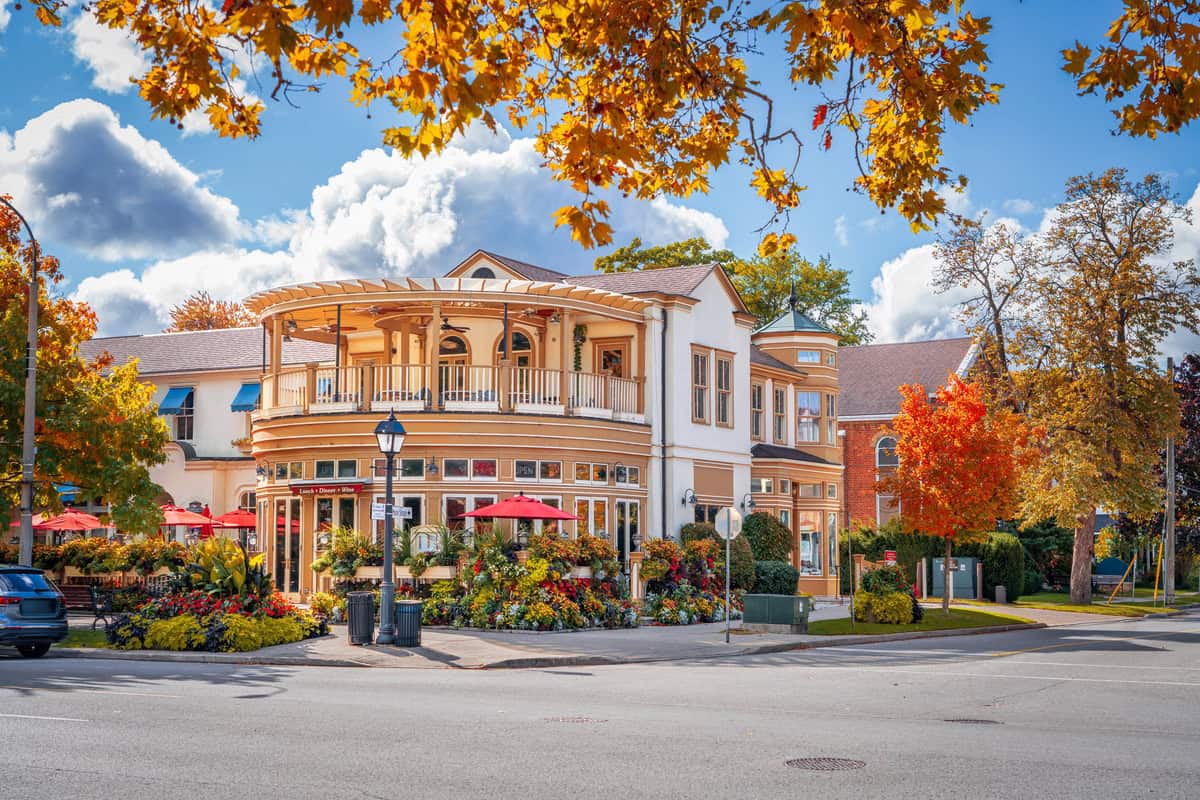 A charming, two-story corner building with a wraparound balcony and outdoor dining patio in Niagara-on-the-Lake Canada is surrounded by vibrant autumn foliage and colorful flower beds. Red umbrellas and blooming plants line the sidewalk café area, creating a cozy, inviting scene under a partly cloudy blue sky in this small Ontario town.