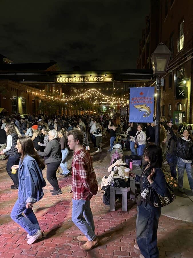 Large crowd gathered at night under twinkle lights at the historic Distillery District, participating in a public line dancing event near the Gooderham & Worts sign.