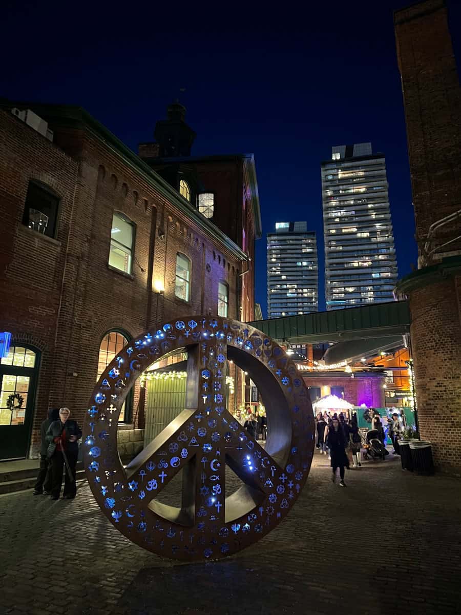 Illuminated peace symbol sculpture adorned with multicultural religious icons in the Distillery District, with historic brick buildings and city towers in the background.
