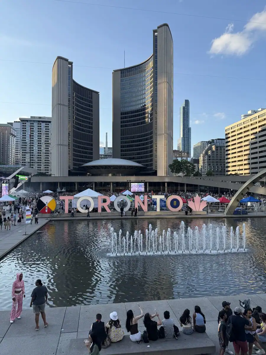 Toronto's iconic sign located at Nathan Phillips Square in downtown Toronto in summer. Behind the sign is Toronto's City Hall and a fountain is in front. Visitors enjoy exploring the area and posing for pictures with the Toronto sign.