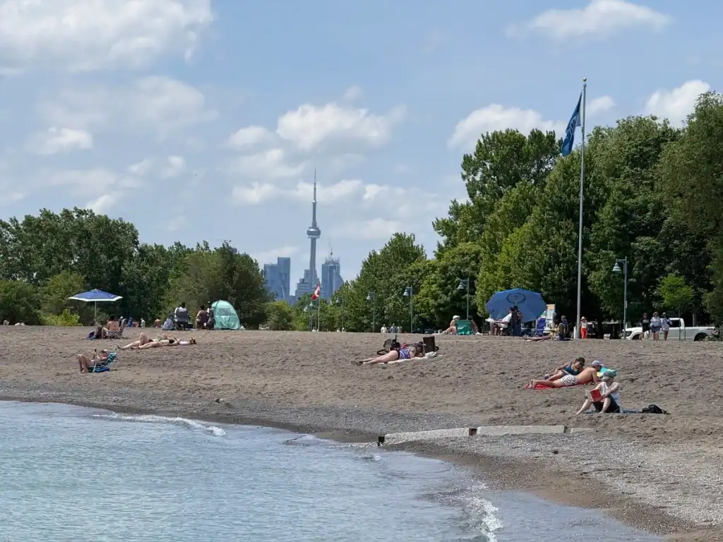 Woodbine Beach with views of the CN Tower on a summer day. People walk the boardwalk and bathers enjoy sitting on the sand along the edge of Lake Ontario.