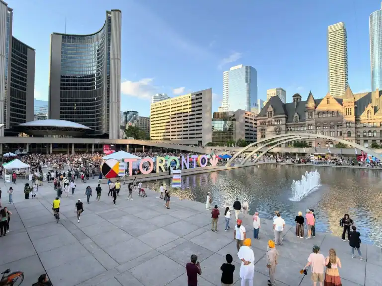 Toronto sign located at Nathan Phillips Square with the fountain in front in summer. Old city hall is also visible and visitors walk around the square in front of Toronto city hall.