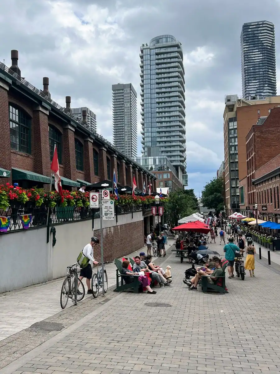 View of Market Street with cyclists, visitors sitting in Muskoka chairs, patios with umbrellas beside the historic St Lawrence Market in downtown Toronto.