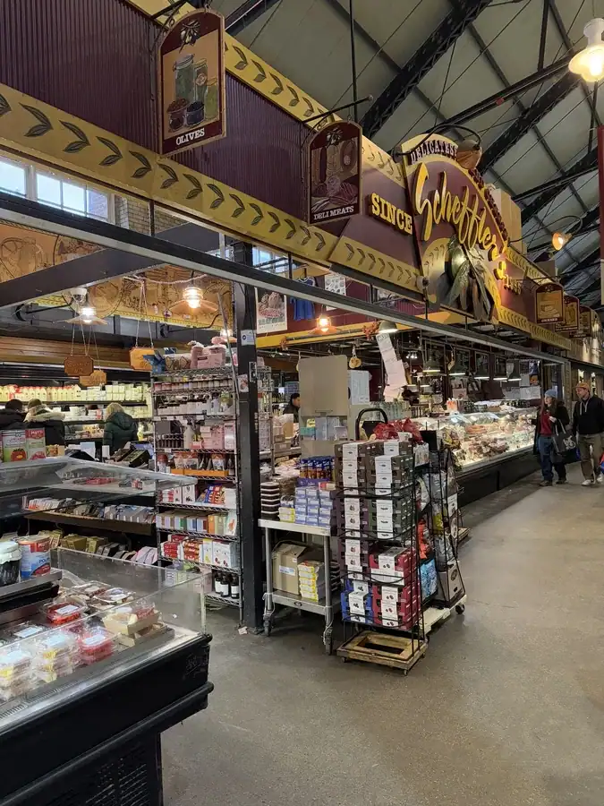 St Lawrence Market cheese vendor with shelves of crackers, cheese, and other displays of food. Visitors walk past the food vendor at the indoor North Market.