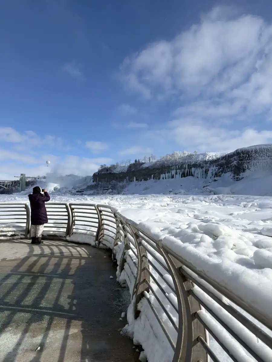 Tourist takes a photo of the frozen Niagara Falls in winter from the observation deck at the Niagara Parks Power Station. The Niagara river is frozen with snow with a bright blue sky above.
