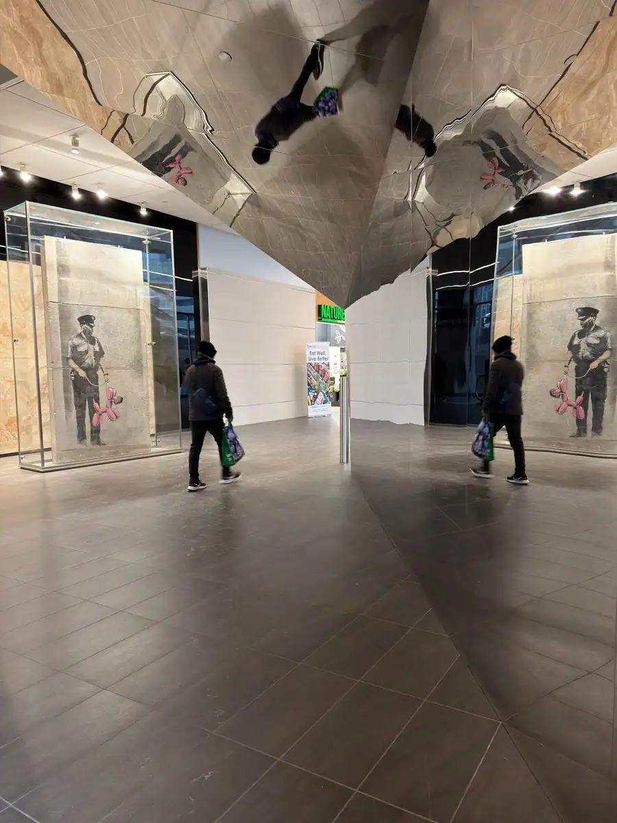 Pedestrians walk past an enclosed Banksy piece of art in Toronto's PATH network. The art is painted on a concrete wall and shows a police officer holding a pink balloon dog.