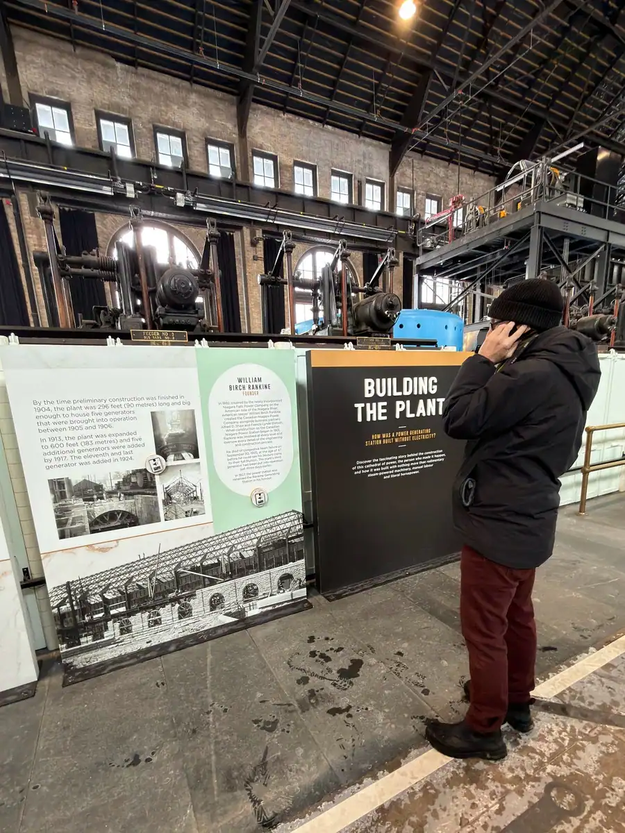 A visitor is standing in front of archival photos of the Niagara Power Station in Niagara Falls Canada. He is holding an audio guide to his ear as he listens to the audio tour of the historic power station.
