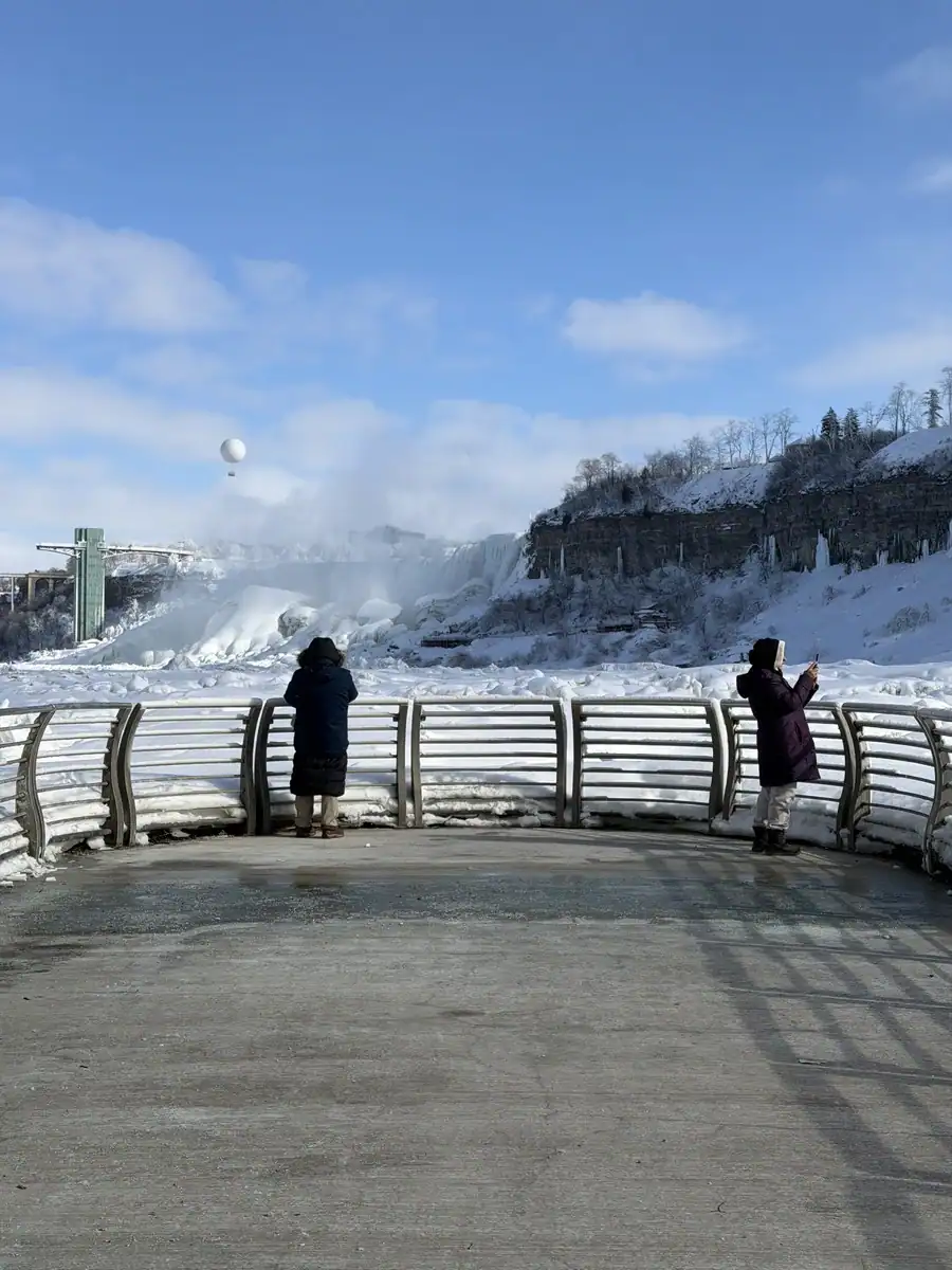 Two visitors stand on the concrete observation deck at the end of the tunnel at the Niagara Power Station in the heart of winter. The frozen Niagara River and American Falls are beyond the fence at the end of the deck.