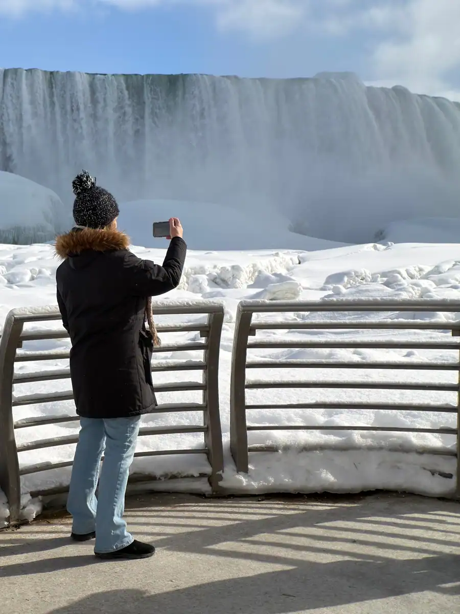 A visitor takes a photo of Niagara Falls in winter from the deck of the observation deck of the Niagara Power Station. Beyond the fence the Niagara River is frozen and the falls are in the distance on a bright, winter day.