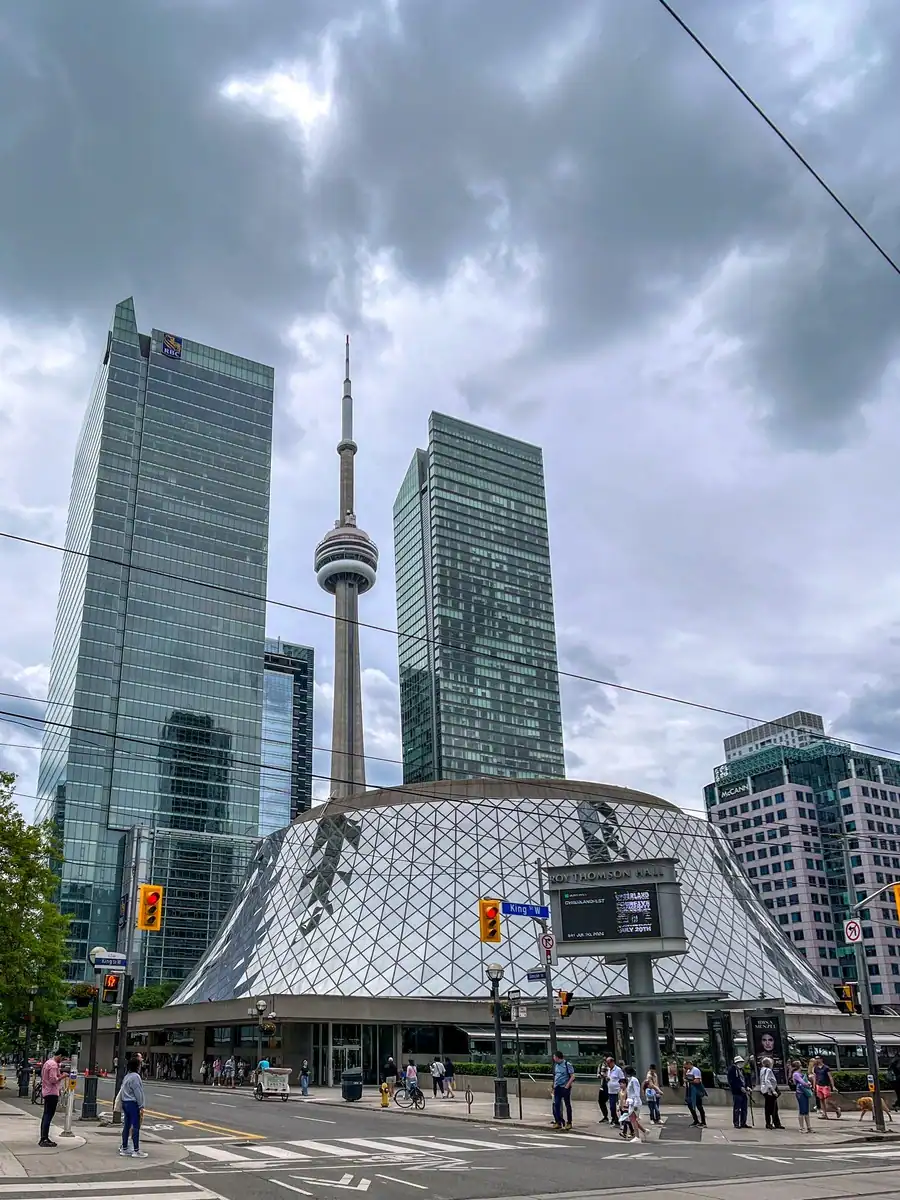 Toronto's Roy Thompson Hall with the CN Tower and skyscraper buildings behind. Pedestrians are walking on the sidewalk in front of the building.