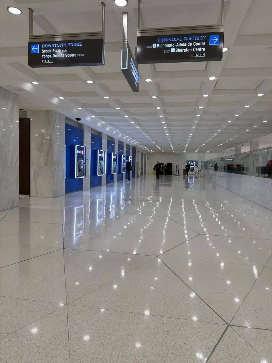 People walk through a brightly lit white underground pathway with white marble as part of Toronto's PATH network. Signs direct pedestrians to various Toronto landmarks.