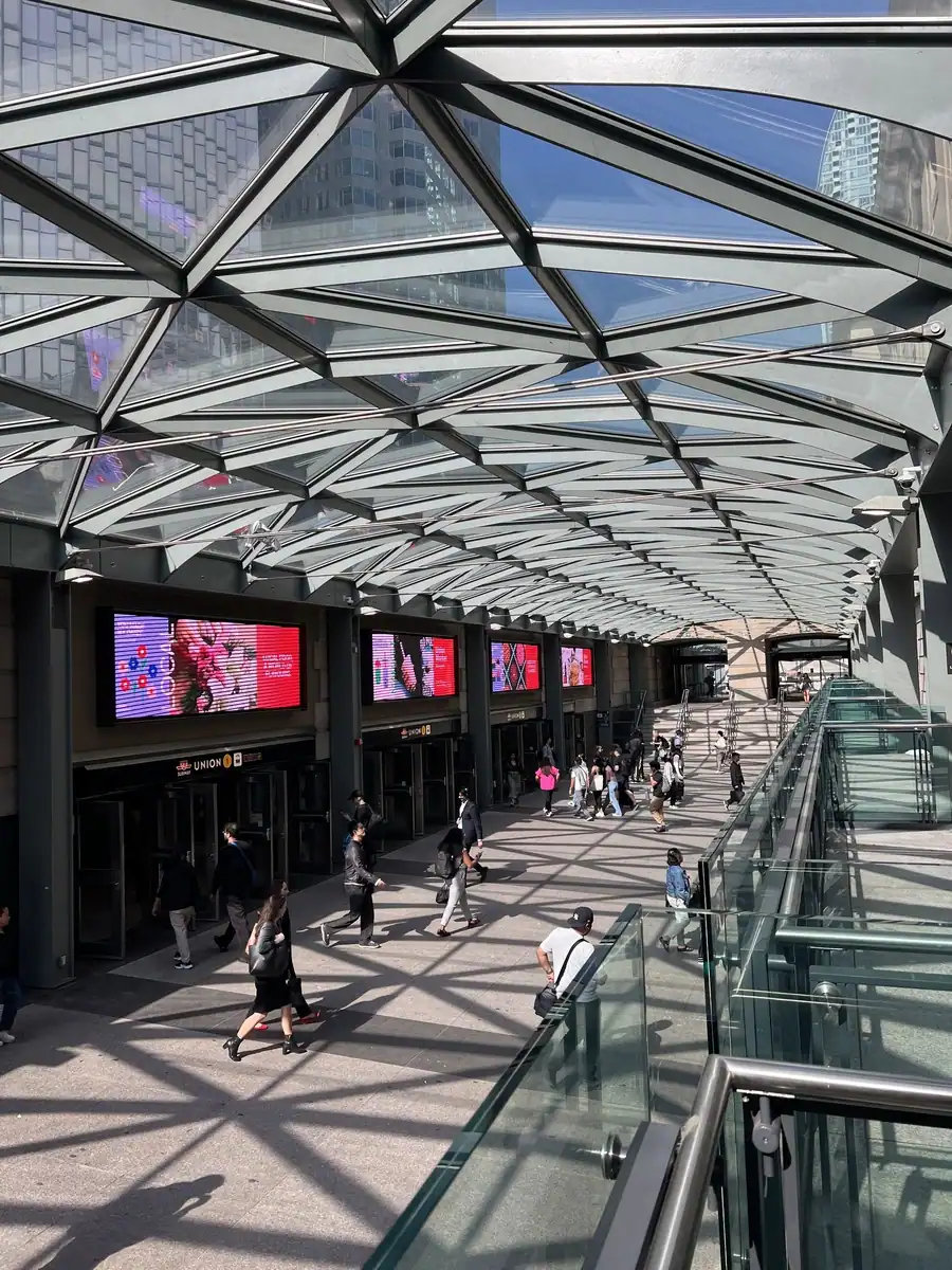 A glass covered walkway connects the trains and subway sections of Union Station in downtown Toronto. People walk under the glass roof on a bright, sunny day.