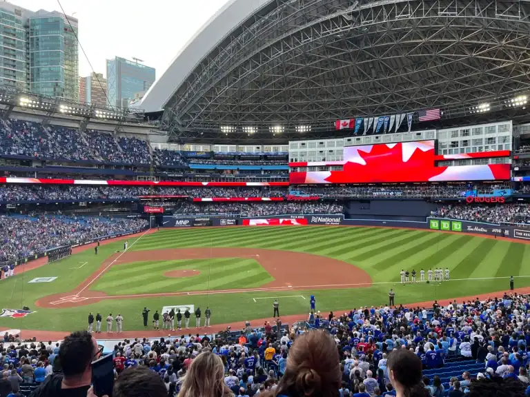 The roof of the Rogers Centre is open on a summer day during a Toronto Blue Jays baseball game. Players are on the field and the Canadian flag is displayed during the national anthem.