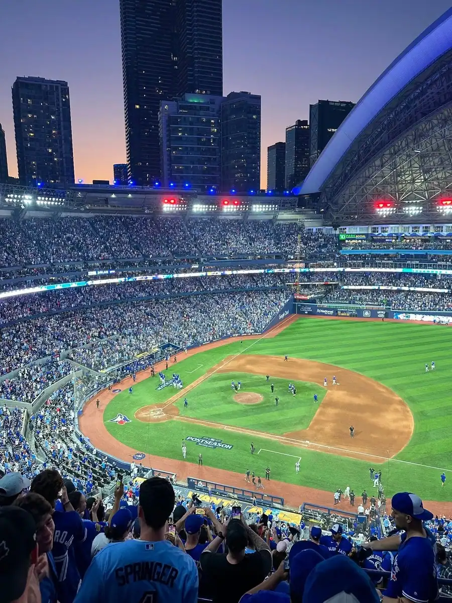 The sun sets over the field of the Rogers Centre on a summer evening with the dome open. Baseball players are on the field and the stands are full of Toronto Blue Jays baseball fans.