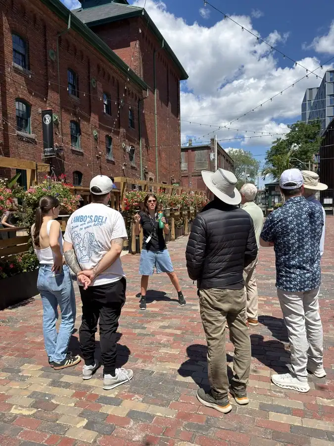 Visitors stand around a tour guide as they explain the history of Toronto's Distillery District during a guided walking tour. Historical buildings are behind the tourists on a sunny summer day.