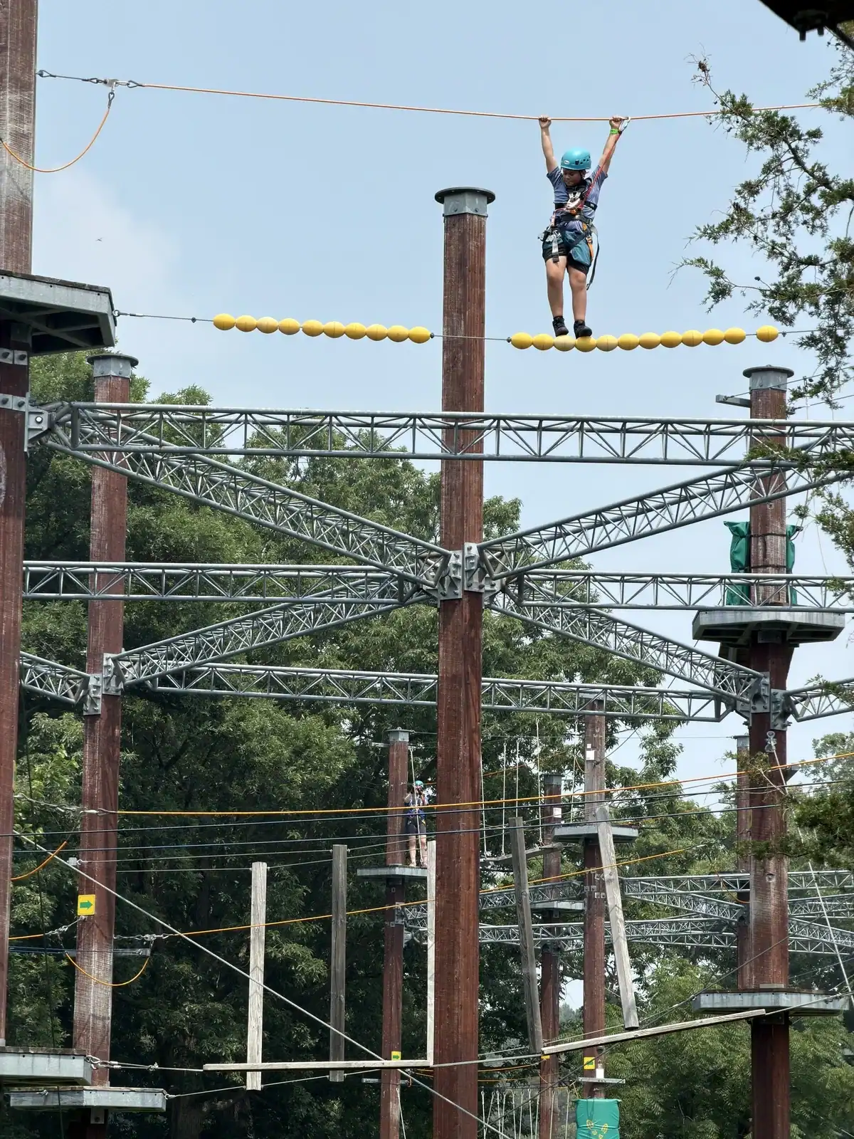 A teenager makes his way across the top of the ropes course on a summer day. He is strapped in with a harness and is wearing a helmet.