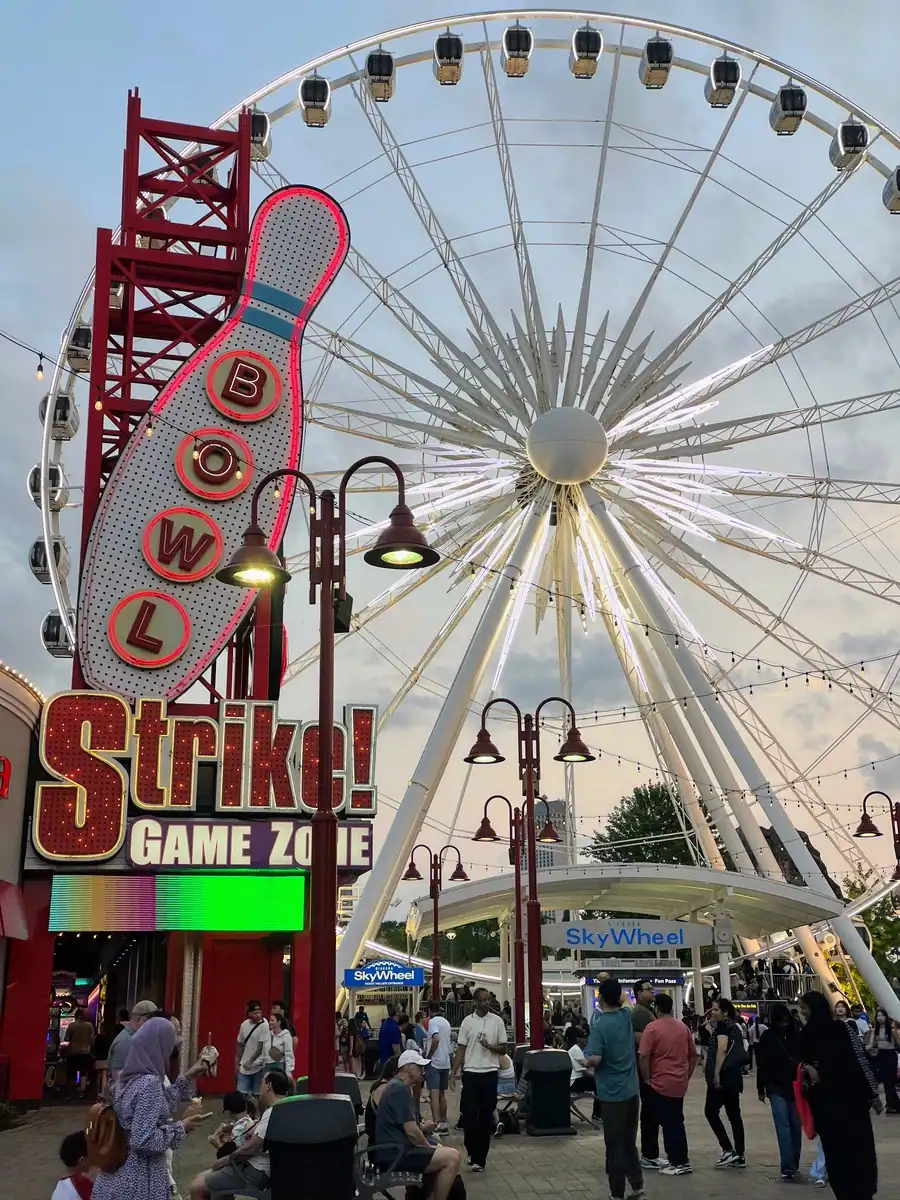 Tourists walk around Clifton Hill in Niagara Falls Canada with the SkyWheel Ferris Wheel towering over the crowds. A neon sign for Stike Bowl is also in front of the ferris wheel.