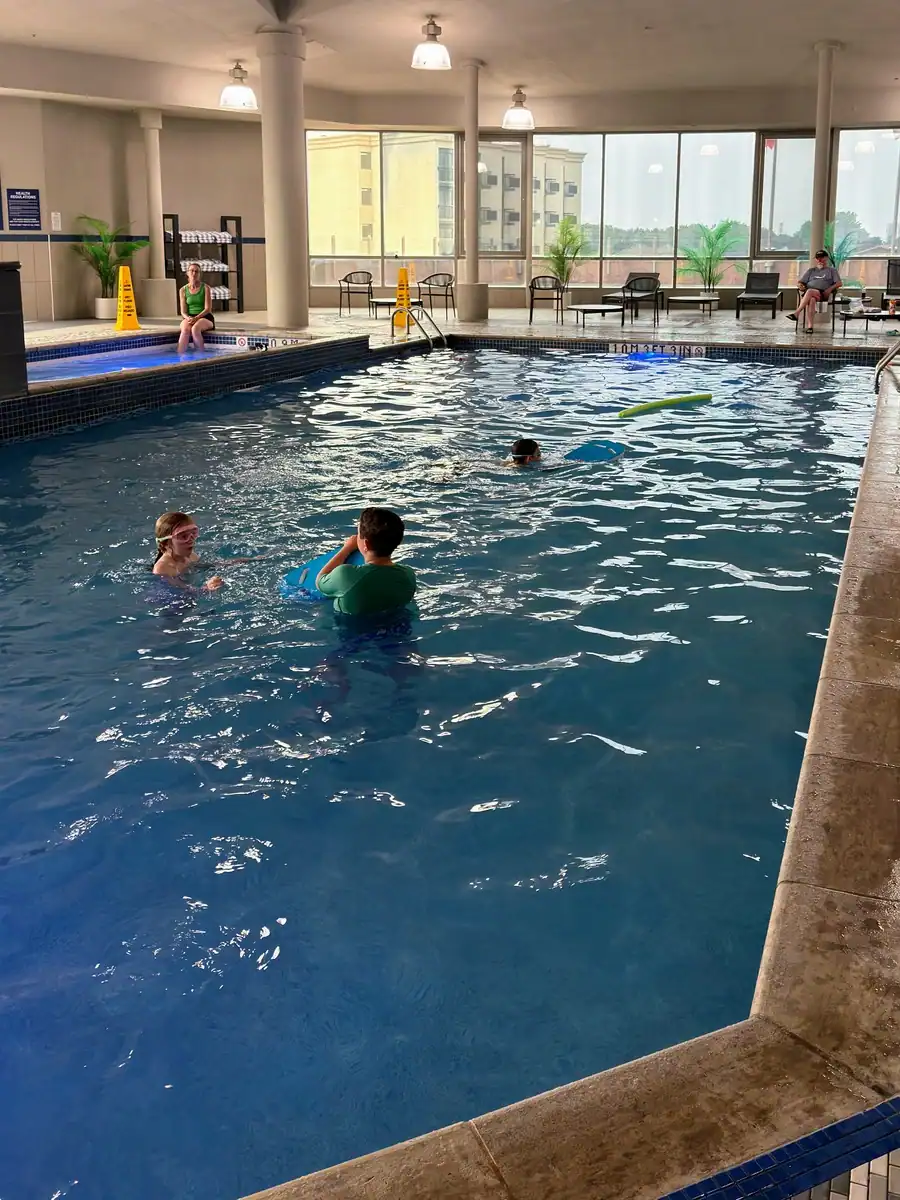 Kids swim in a large indoor pool at the Hyatt Regency in Niagara Falls Canada. Windows overlooking the city, and a large hot tub are also located beside the large pool.