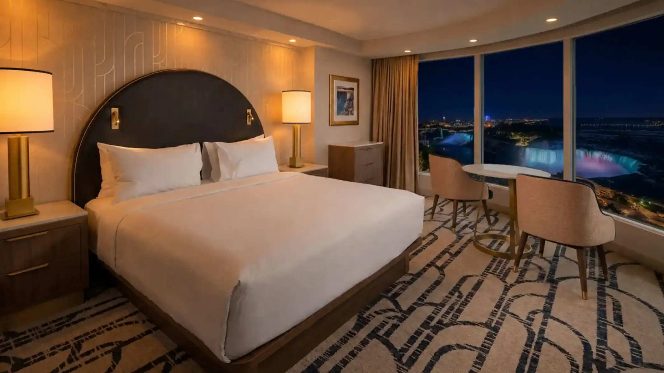 Hotel room with King bed and small table with two chairs in front of large floor to ceiling windows with views of the illuminated Niagara Falls at night.